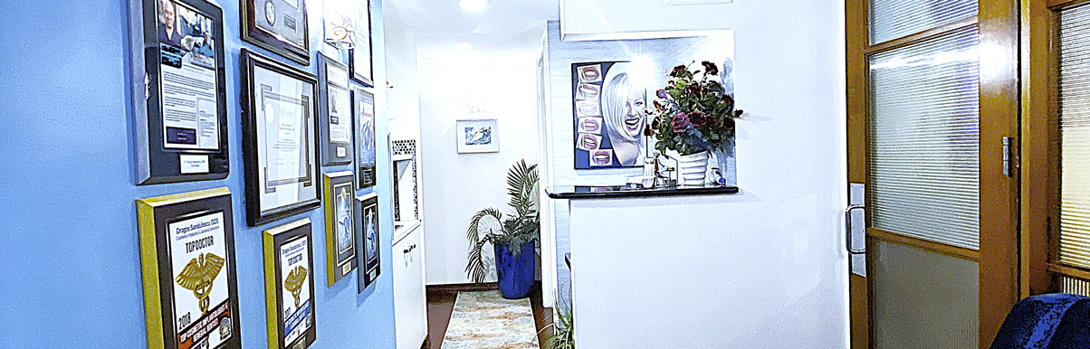 fifthavenuesmile | Hallway with blue accent wall displaying framed dental awards and certificates, decorative plants, and artwork of a smiling face.